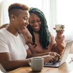 Female couple looks at a laptop together, smiling and drinking coffee.